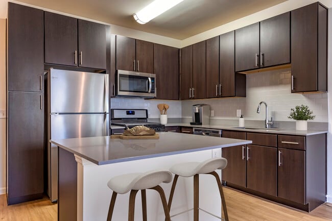Newly renovated apartment homes feature kitchens with espresso cabinetry, grey quartz countertops, stainless steel appliances, grey tile backsplash, and hard surface flooring