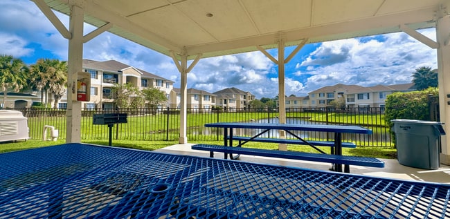 Serene apartment view from a covered patio with blue picnic tables, a barbecue grill, and lush greenery around a tranquil pond. - The Village at Melbourne