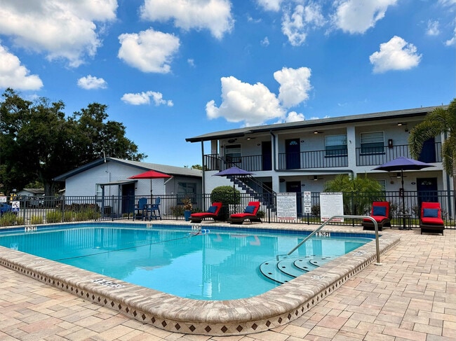 Charming apartment complex with a sparkling pool surrounded by sun loungers, vibrant umbrellas, and lush greenery under a bright blue sky. - Waterside Village
