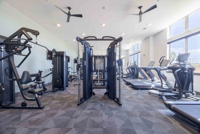 Las máquinas de entrenamiento funcional con cables y las máquinas para piernas son los elementos centrales del área de entrenamiento de fuerza del gimnasio Garrett Parker, con equipos cardiovasculares visibles cerca de las ventanas.