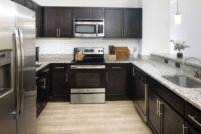 Renovated Package II Kitchen with dark cabinetry, speckled granite countertops, white tile backsplash, stainless steel appliances, pendant lighting, and hard surface flooring - AVA South End Apartments