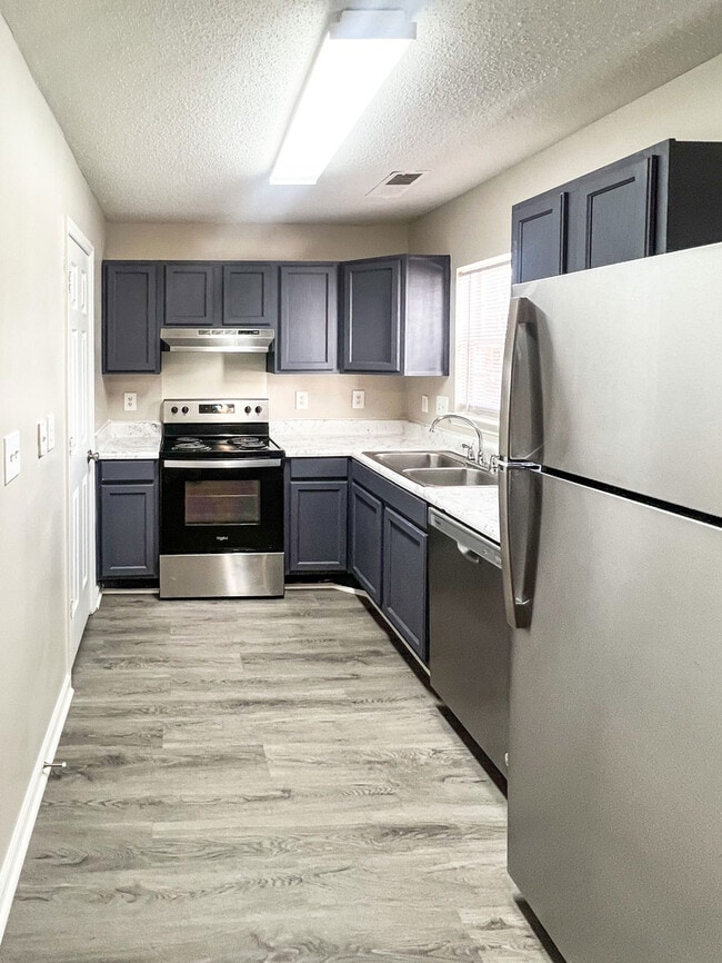 Galley kitchen view with stainless steel refrigerator, dishwasher, dark cabinetry, marble style counters, and wood look flooring - Pendleton Townhomes