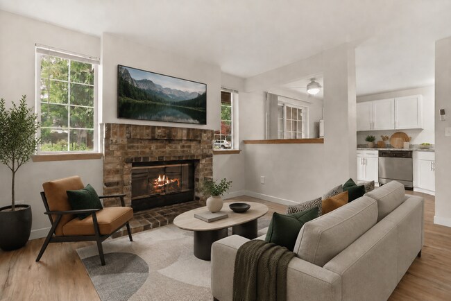 Staged apartment living room with a brick fireplace, cream seating, scenic TV, and an adjacent kitchen with stainless-steel appliances.