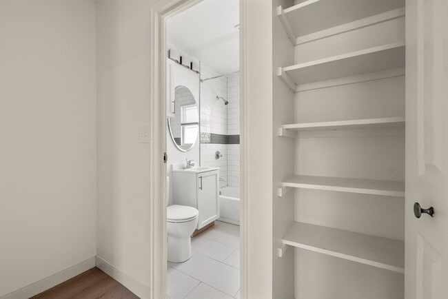Modern bathroom with sleek white tiles, a stylish round mirror, and an adjacent open shelving unit for ample storage. - Stony Brook Commons