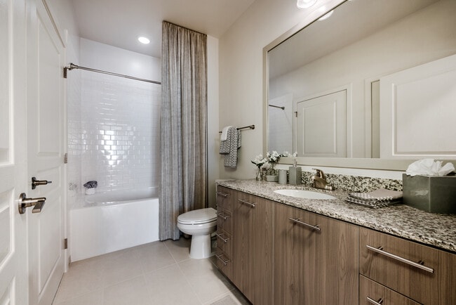 Baño con encimeras de granito, gabinetes de color marrón, pisos de superficie dura y bañera con azulejos - Avalon Arundel Crossing