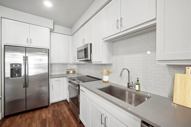 Cocina con acabados de segunda calidad, que incluye electrodomésticos de acero inoxidable, gabinetes blancos, salpicadero de azulejos blancos y encimeras de cuarzo gris.