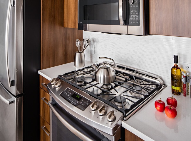 Signature Collection Kitchen with oak cabinetry, light grey quartz countertops, white tile backsplash, upgraded stainless steel appliances, and hard surface plank flooring