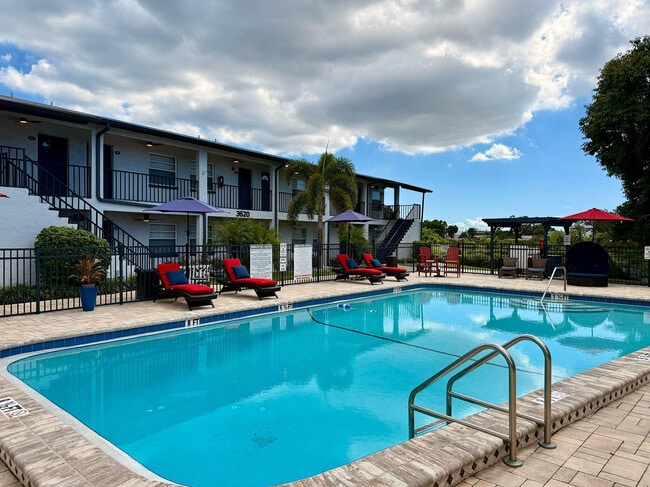 Inviting apartment complex with a sparkling pool, cozy red loungers, and a charming outdoor seating area under umbrellas.