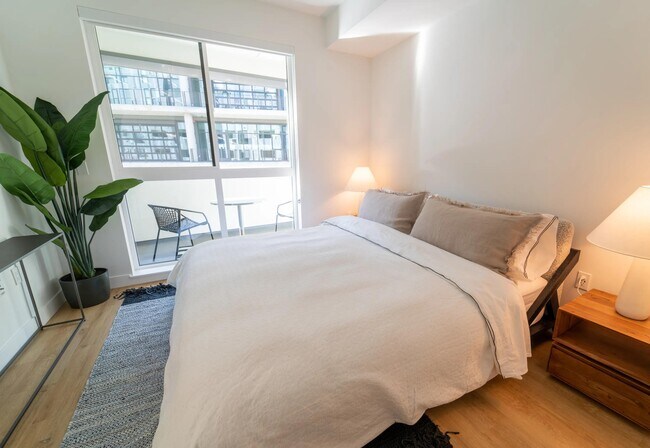 Bedroom with a private balcony, warm wood-style plank flooring, and a bright, calming atmosphere shaped by abundant natural light.