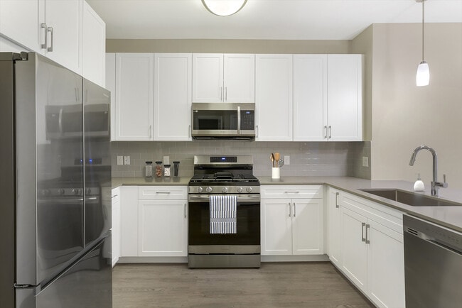 Renovated Package II kitchen with grey quartz countertops, white cabinetry, grey tile backsplash, stainless steel appliances, and hard surface flooring