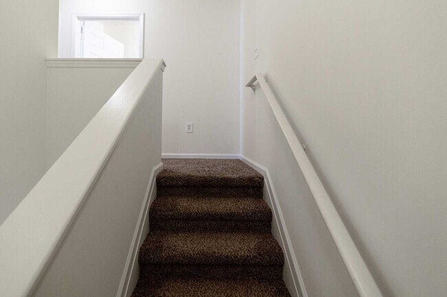Carpeted staircase with white railings and walls leading to an upper hallway area with an open doorway and wall outlet