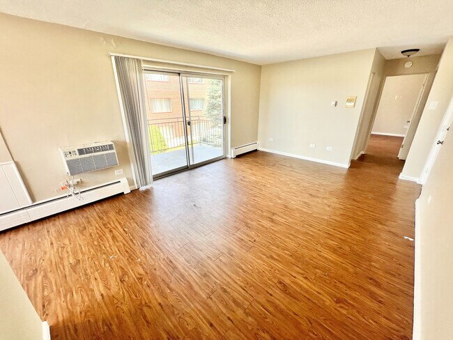 Luminosa sala de estar con elegantes pisos de madera, grandes puertas corredizas de vidrio y moderna unidad de aire acondicionado montada en la pared, que conduce a un acogedor balcón. - Bonneville Apts