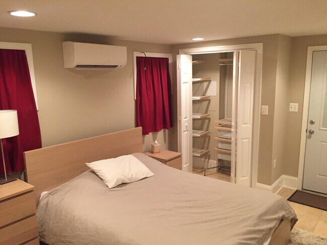 Spacious bedroom with secure windows, secure exterior door, two large closets, including one with Elfa system, large dresser, nightstand, rug, lamps, Minisplit HVAC unit, and mirror.