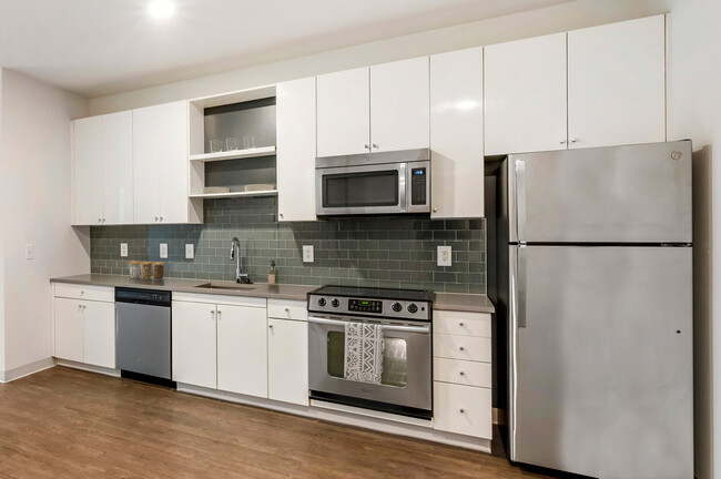 Renovated Package I kitchen with stainless steel appliances, grey quartz countertops, white cabinetry, grey tile backsplash, and hard surface flooring - AVA H Street Apartments