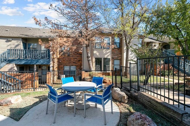 Outdoor courtyard seating area with round table, blue chairs, black metal fence, mature trees, and apartment buildings